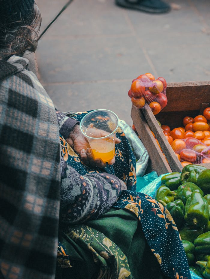 A street vendor holding a juice cup next to crates of fresh produce, including peppers and tomatoes.